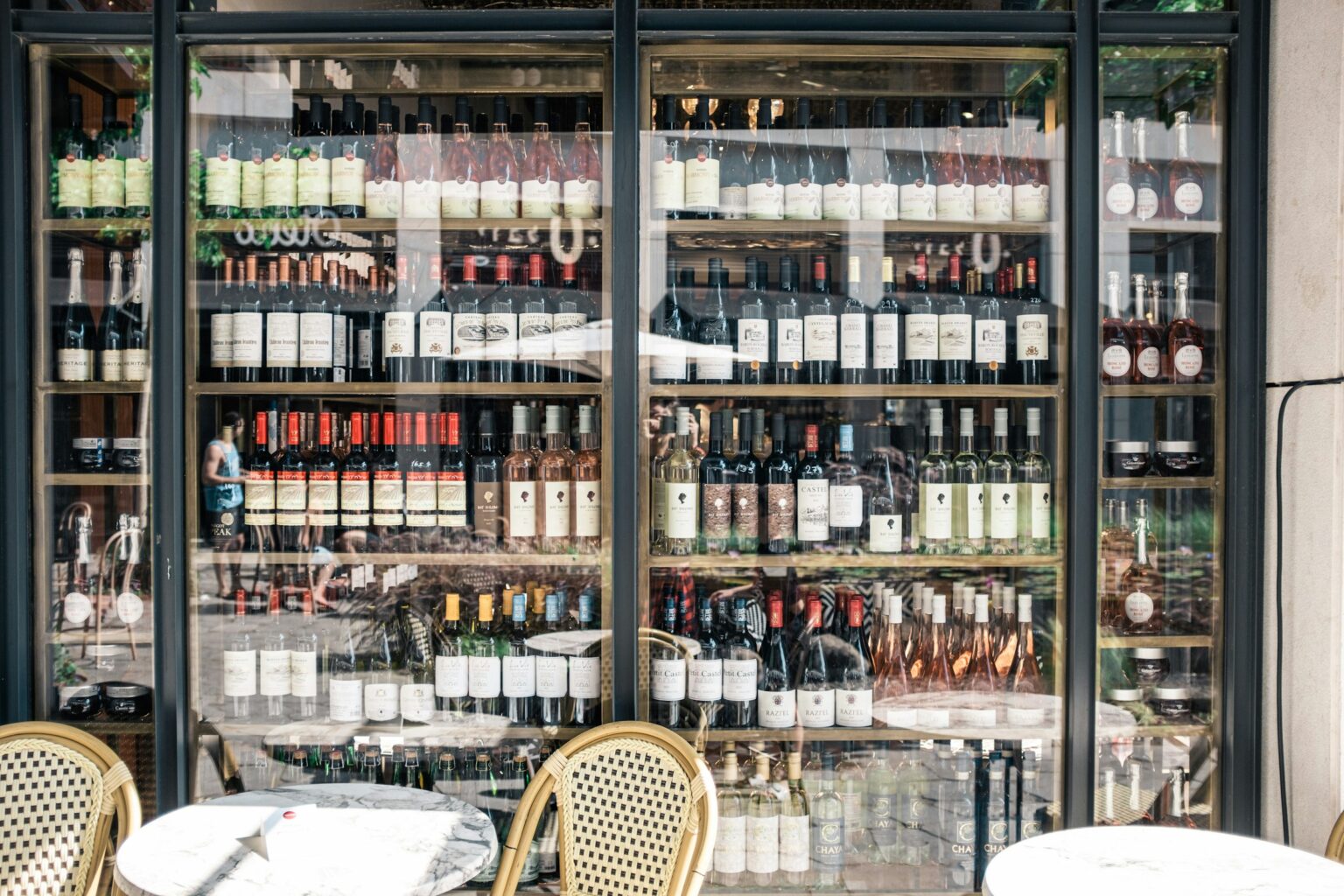 The Importer Tier: How A Wine’s Path To Your Shelf Decides Its Identity A glass-fronted wine shop display photographed through the storefront window, with sidewalk café chairs in the foreground