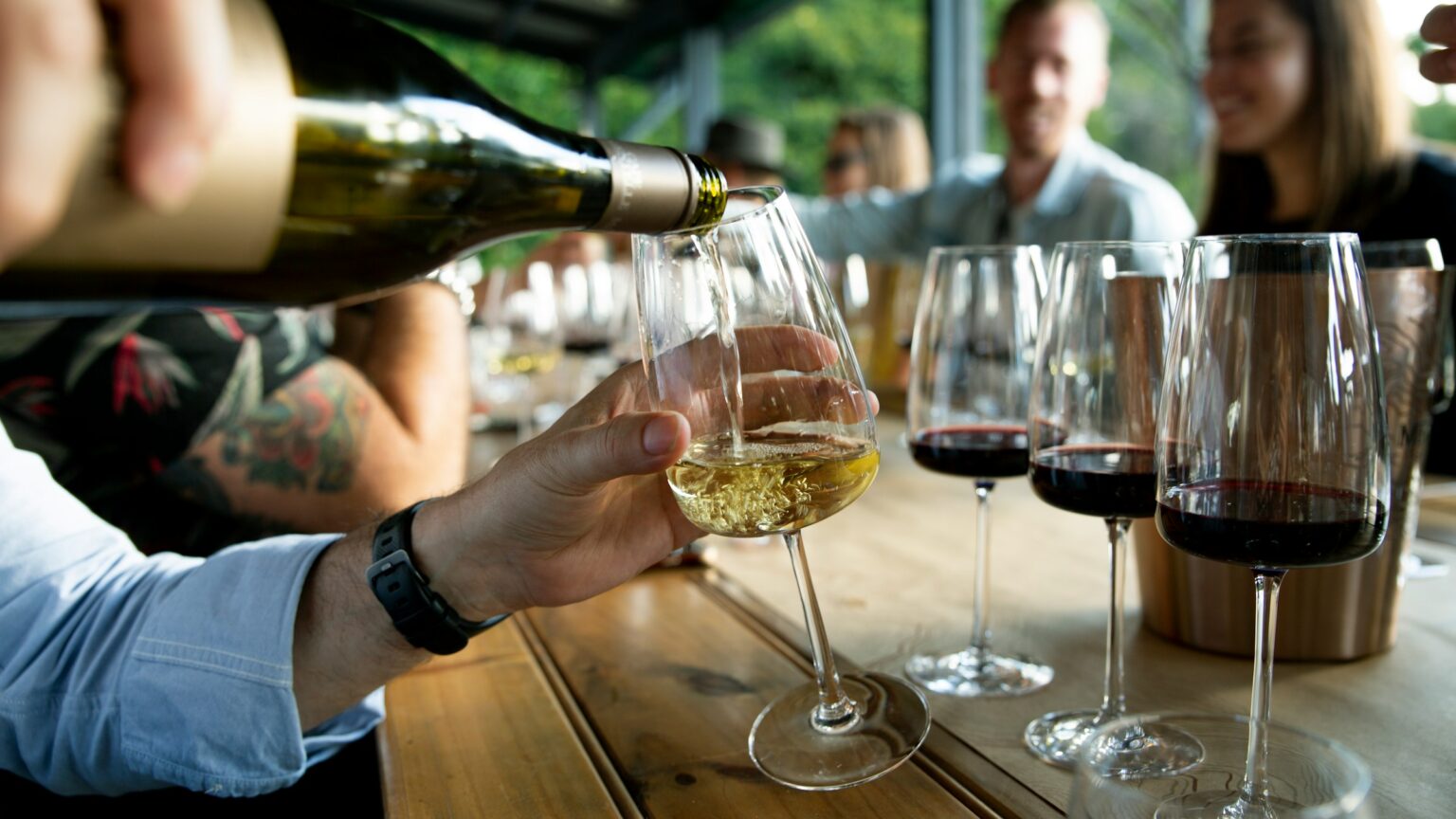 The Corkage Question: What It Costs To Bring Your Own Wine, And What It Buys You Tableside wine service with a bottle being poured into a glass, illustrating the corkage question of what bringing your own wine to a restaurant actually costs and what the fee buys in service and social contract