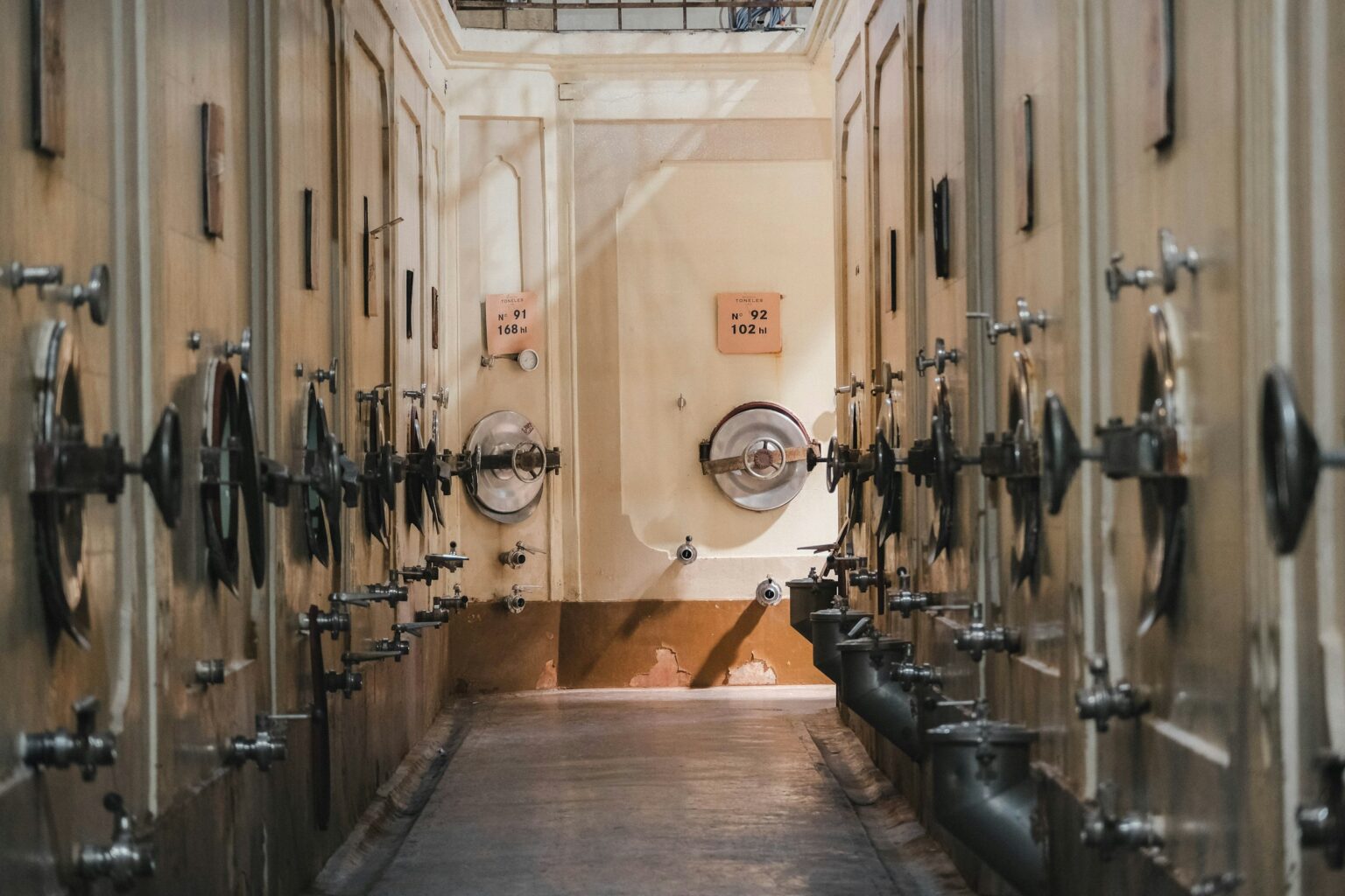 Native Yeast Vs Cultured: The Fermentation Decision That Shapes Wine’s Most Argued Question A winery fermentation hall lined with numbered stainless-steel tank doors and pressure gauges, where the native-versus-cultured yeast decision is made.