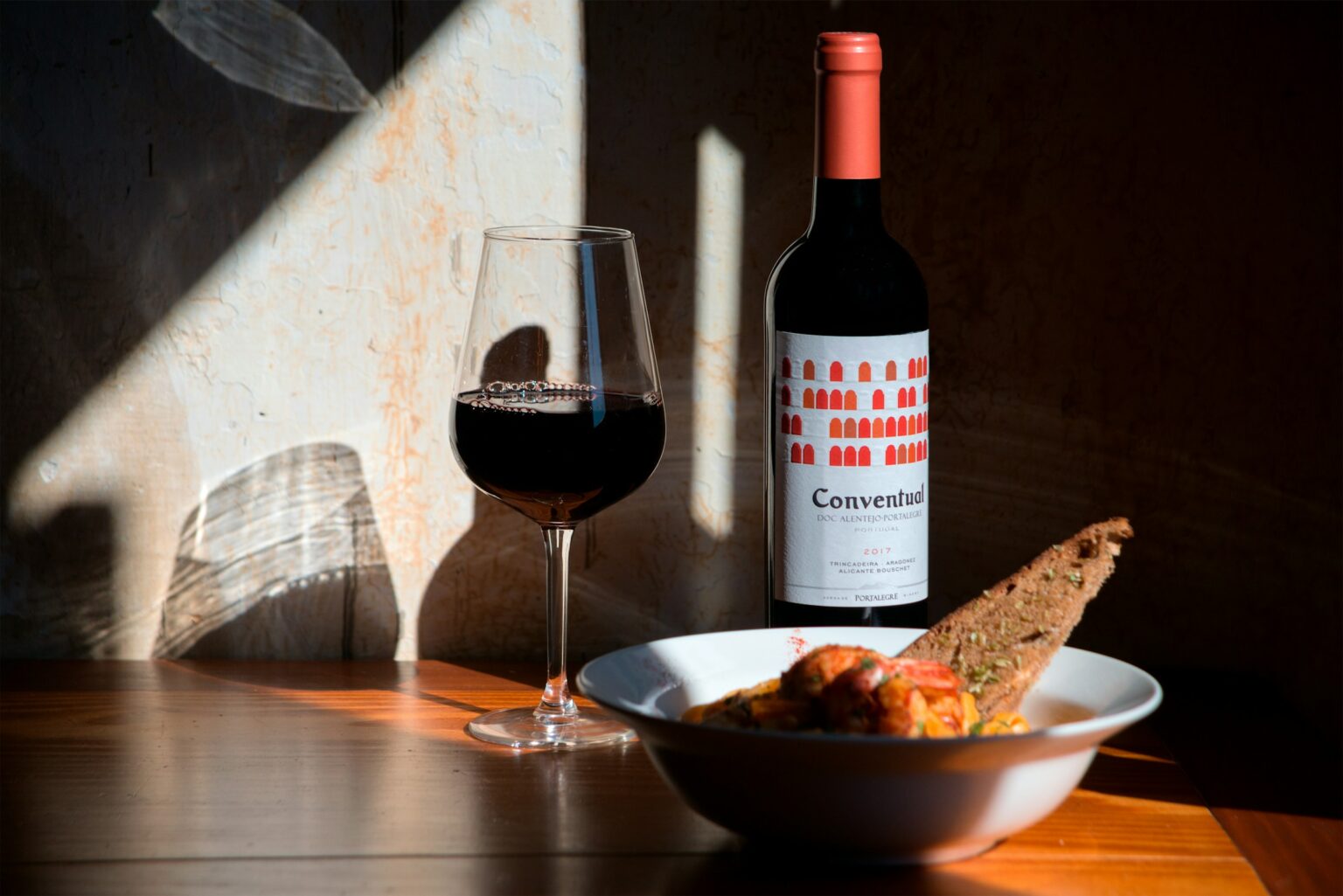 The Heat Equation: Why Your Wine Makes Spicy Food Burn Hotter (And What to Pour Instead) A glass of red wine and an open bottle beside a warm, sauce-heavy dish in raked window light, the high-alcohol red that intensifies chili heat rather than relieving it.
